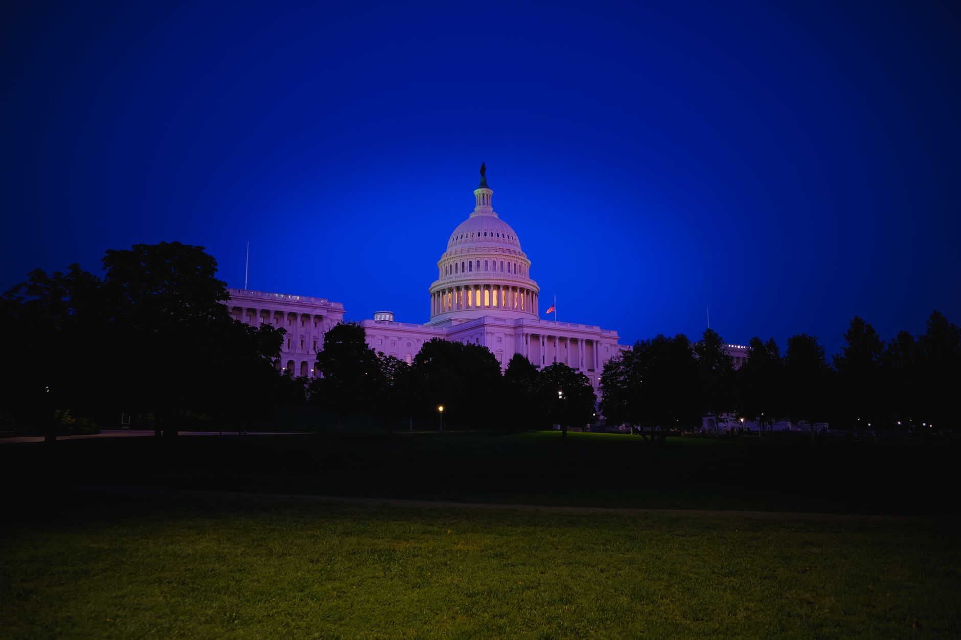 Grand bâtiment néoclassique à dôme illuminé au crépuscule, derrière des arbres et une pelouse sombre, sous un ciel bleu profond.