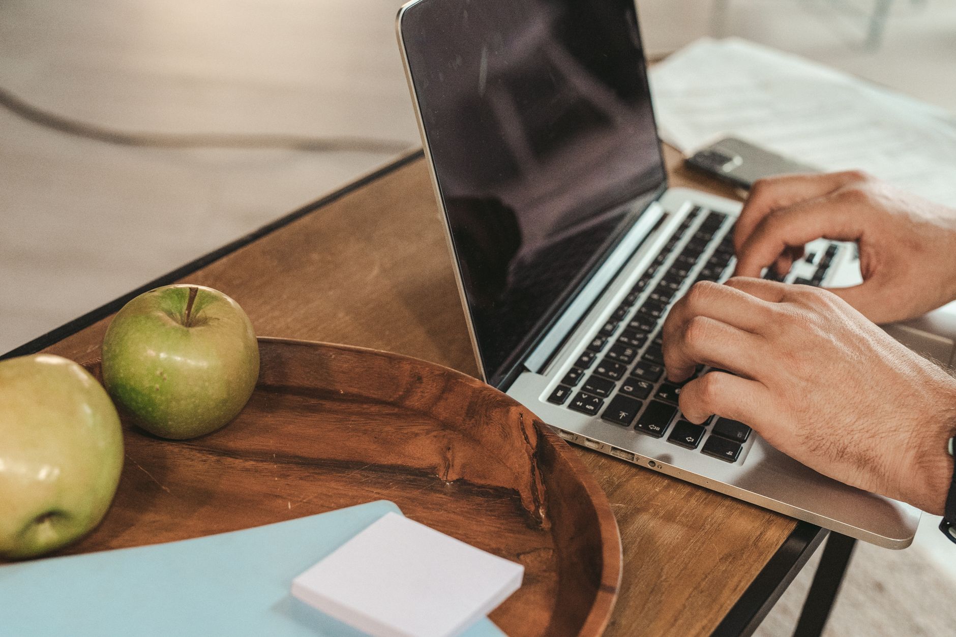 Mains tapant sur un ordinateur portable posé sur un bureau en bois, avec des pommes vertes, un carnet bleu et un bloc de notes au premier plan.