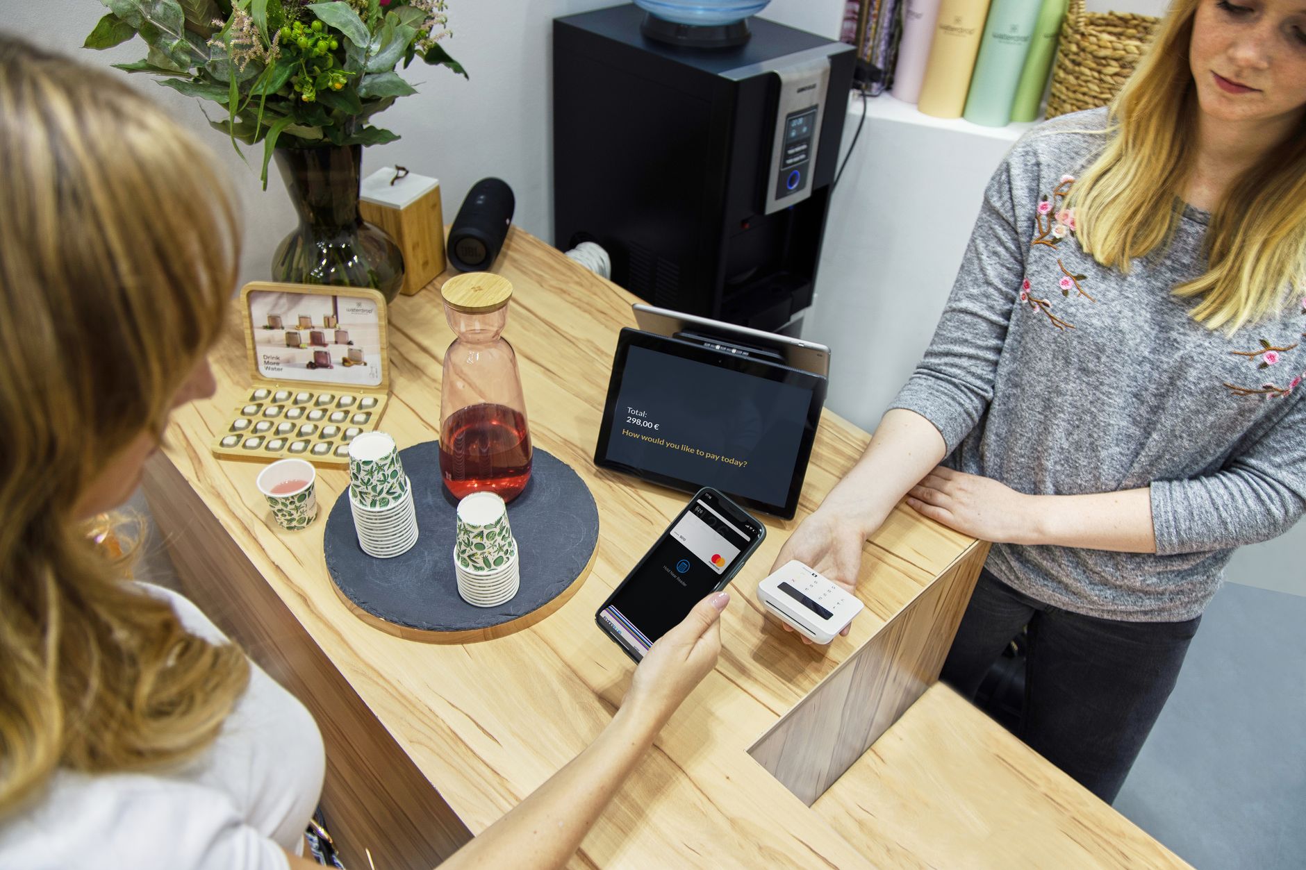 Une cliente règle un achat avec son smartphone près d’un terminal de paiement, devant une vendeuse et une tablette de caisse au comptoir.
